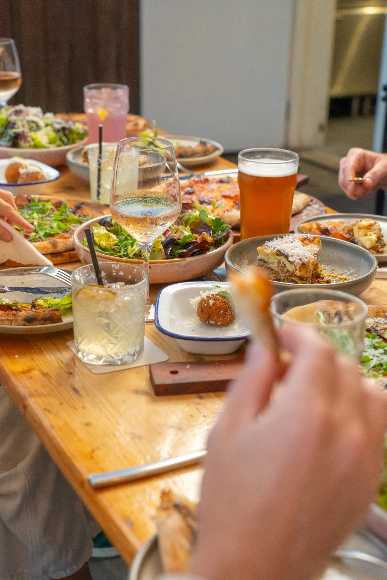Long shared table of pizza, salads and arancini with guests eating and drinking